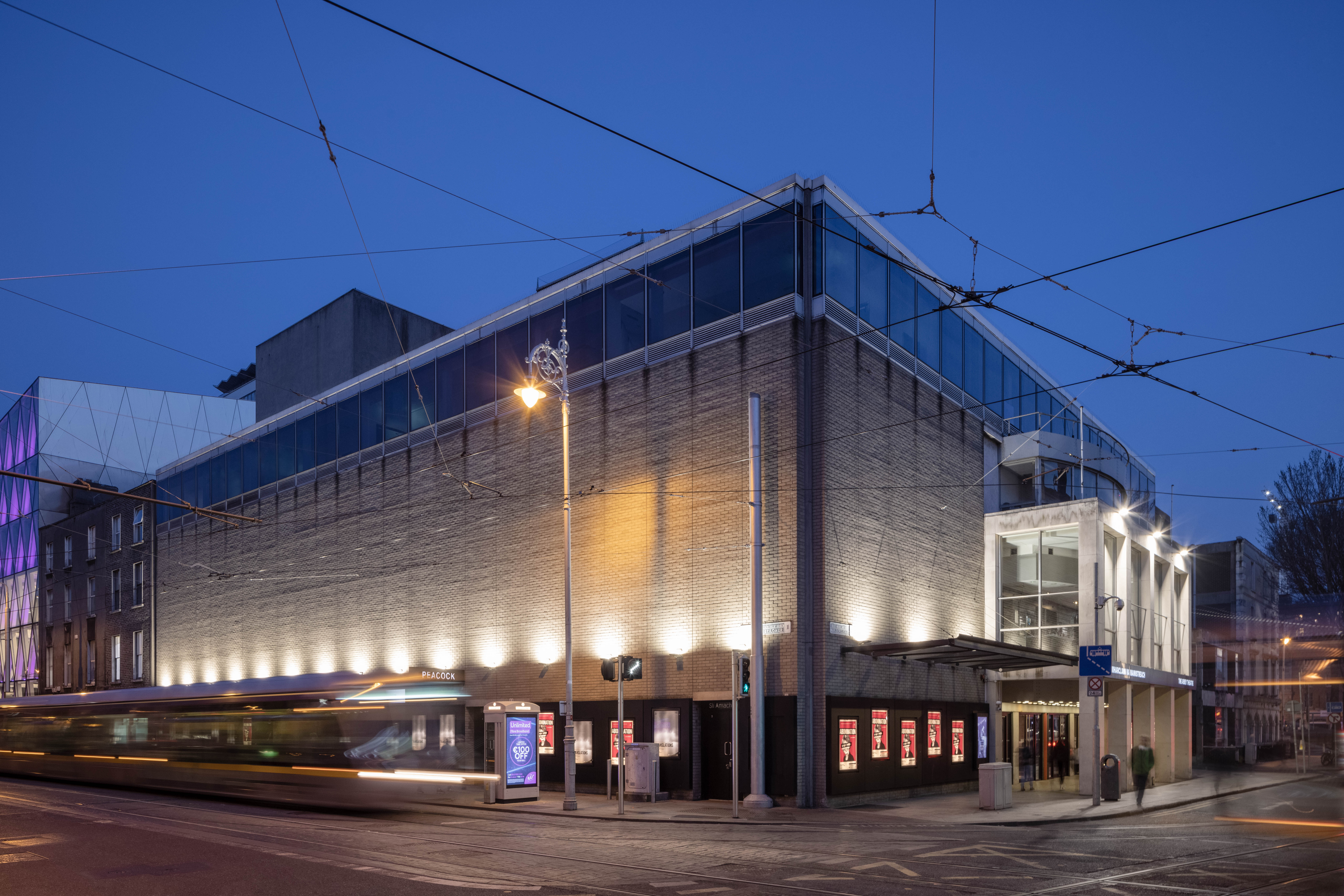An external view of the Abbey Theatre. Photographed from across the street, it shows both the main entrance and glass portico on Marlborough Street, and the entrance to the Peacock Theatre on Abbey Street. The LUAS tram is passing by on Abbey Street, and the image is taken at dusk.