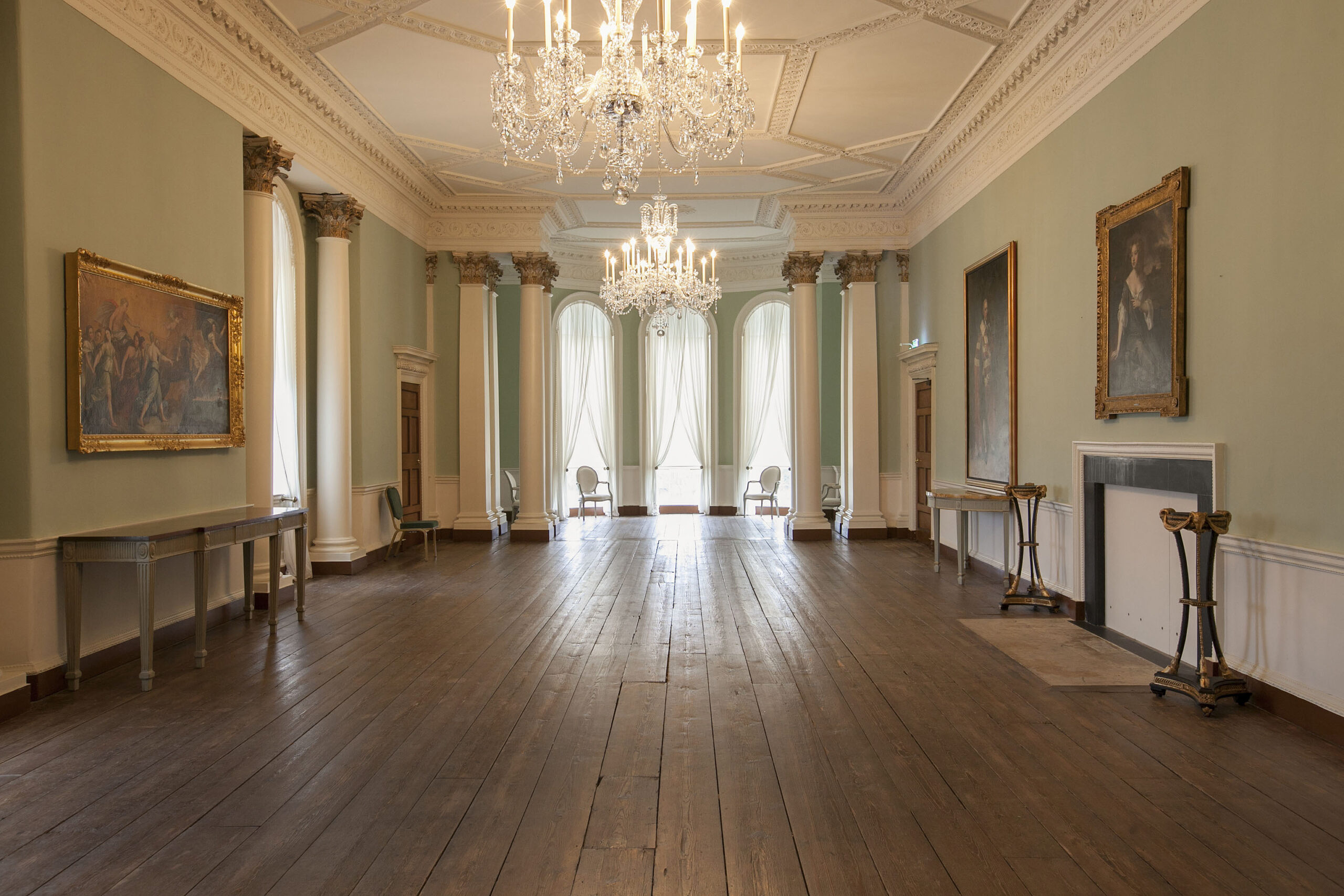 The Ballroom. A long room with arched windows in a bow area at the end of the room. There is a polished wooden floor, there are two ornate chandeliers and paintings on either side of the room. Two columns with ornate capitels flank the bow area.