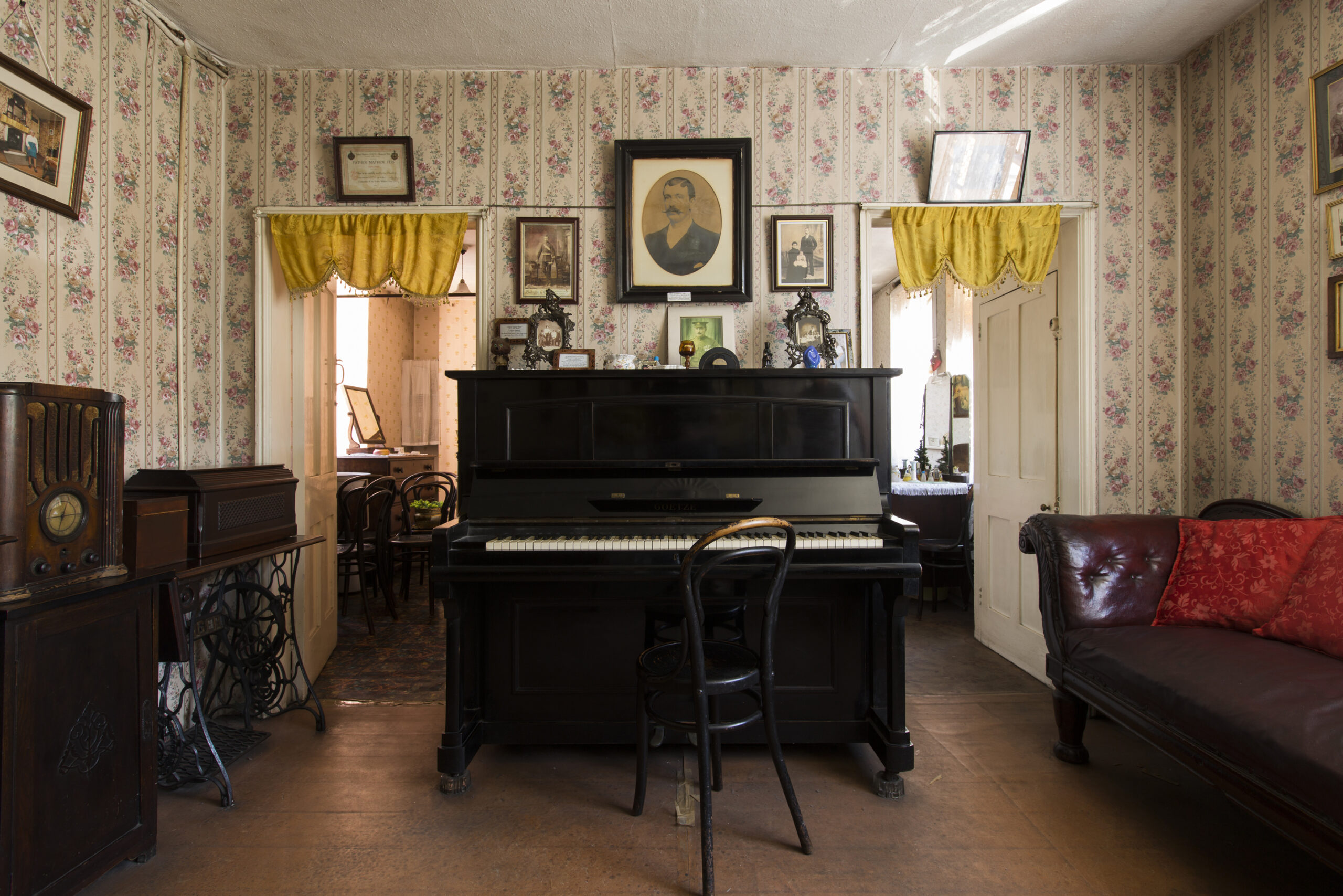 Interior view of living room with upright piano on the centre. The walls are covered with wallpaper with yellow flowers and portraits