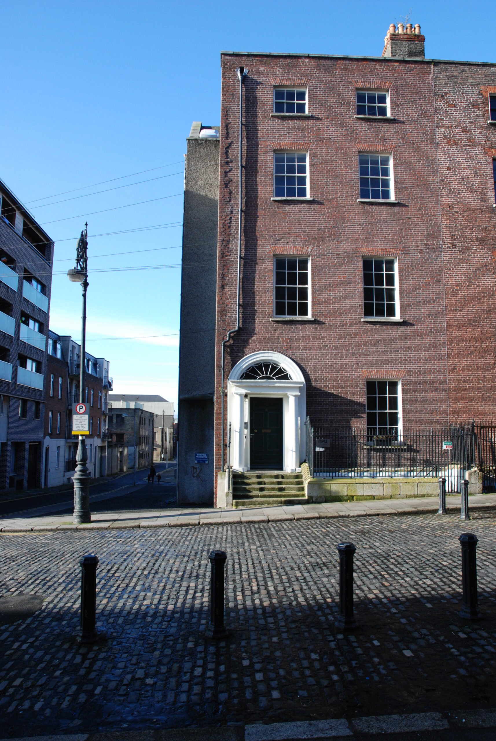 View across cobbled street to a four storey red-brick building with granite steps to front door, and a side-street to the left-hand side.