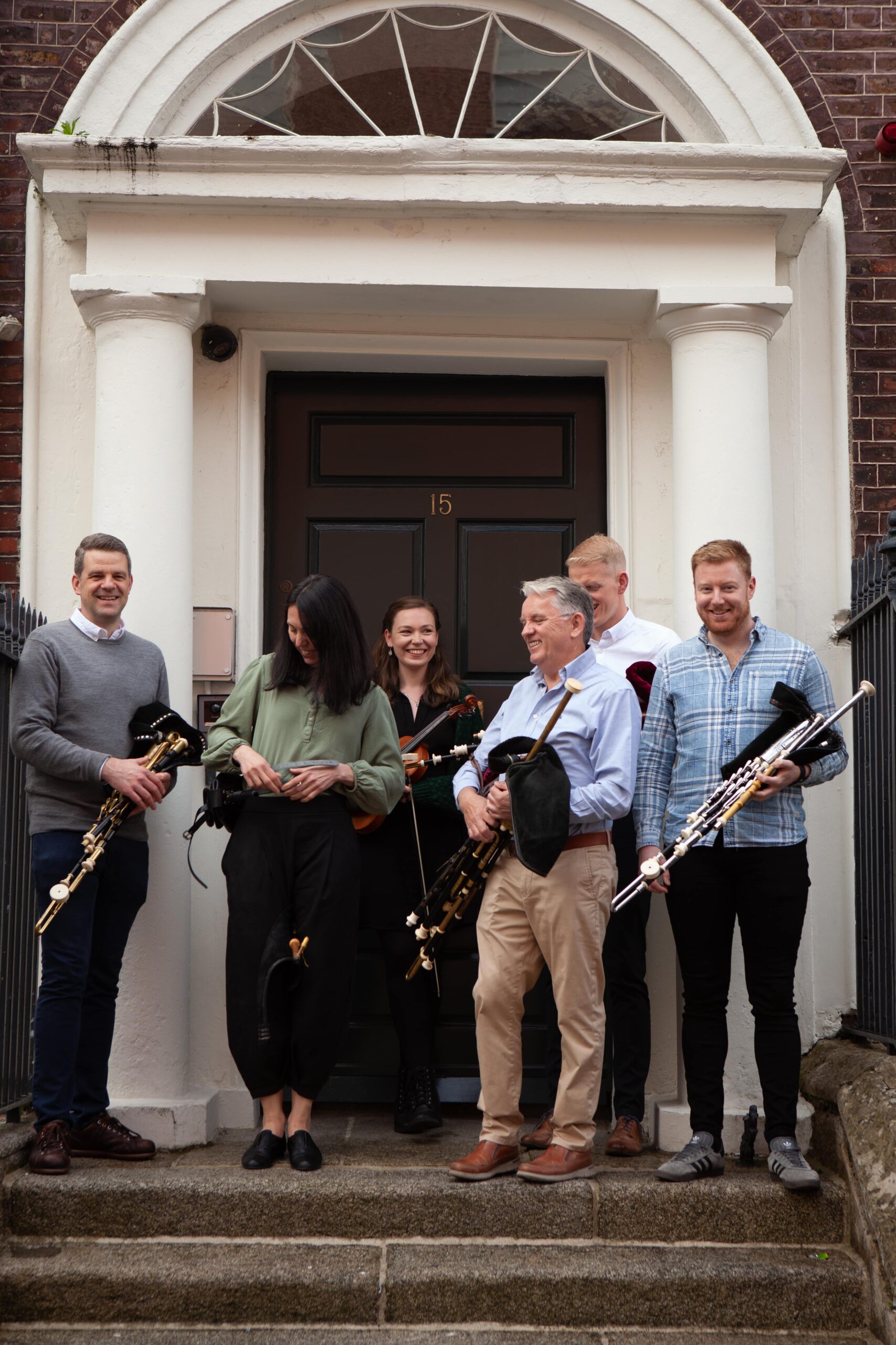 Uilleann pipers stand on steps of a Georgian building, outside the front door, holding their instruments.