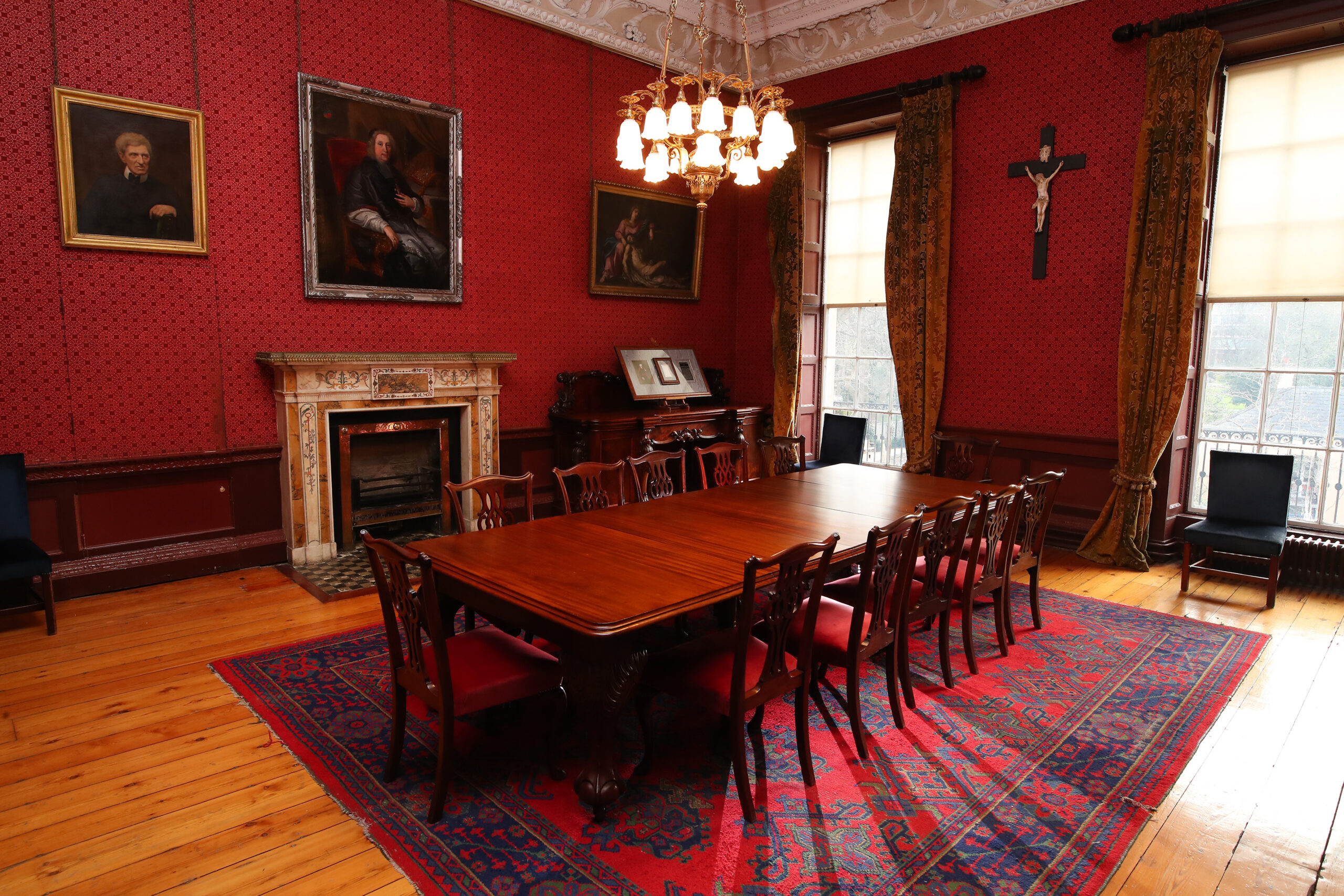 A small, dark red room with a mahogany table in the centre, placed on a dark red and blue carpet and under a large chandelier.