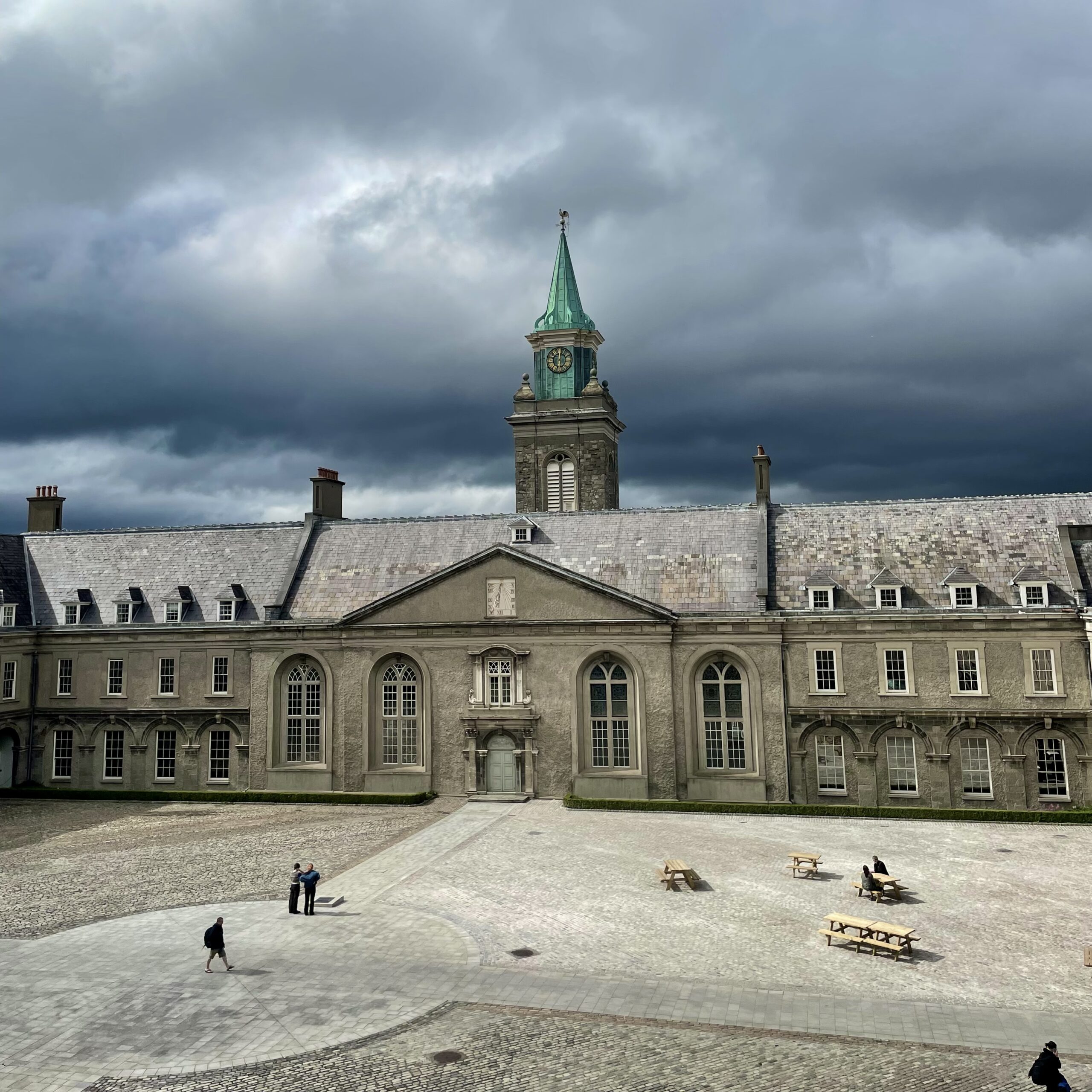 A grey historic building with large arch windows and central grey door, a clock tower with green spire in the centre of the roof, overlooking a cobbled courtyard with a few picnic-style seat tables and some people walking and sitting. The sky is brooding, cloudy and grey.