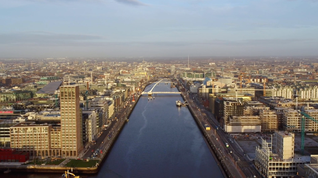 An aerial view of Dublin city looking directly down the river with buidlings on either side. A white bridge, the Samuel Beckett bridge, can be seen in the distance and then a hazy sky as a backdrop to the cityscape.