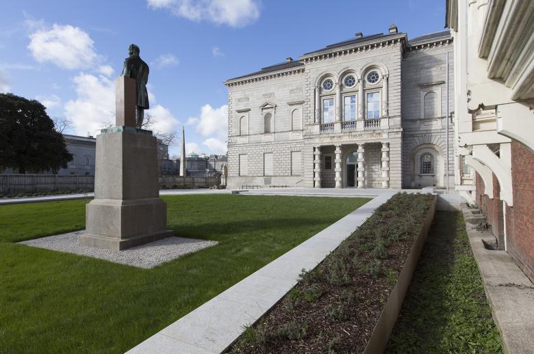 A view of the front of the National gallery of Ireland. The back of a statue is in the left foreground. A lawn leads up to the pillared entrance. There are blue skies with white fluffy clouds.