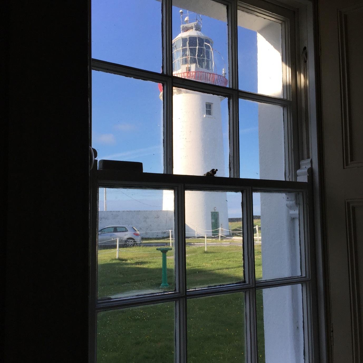 Looking through a window to a white light house surrounded by green grass